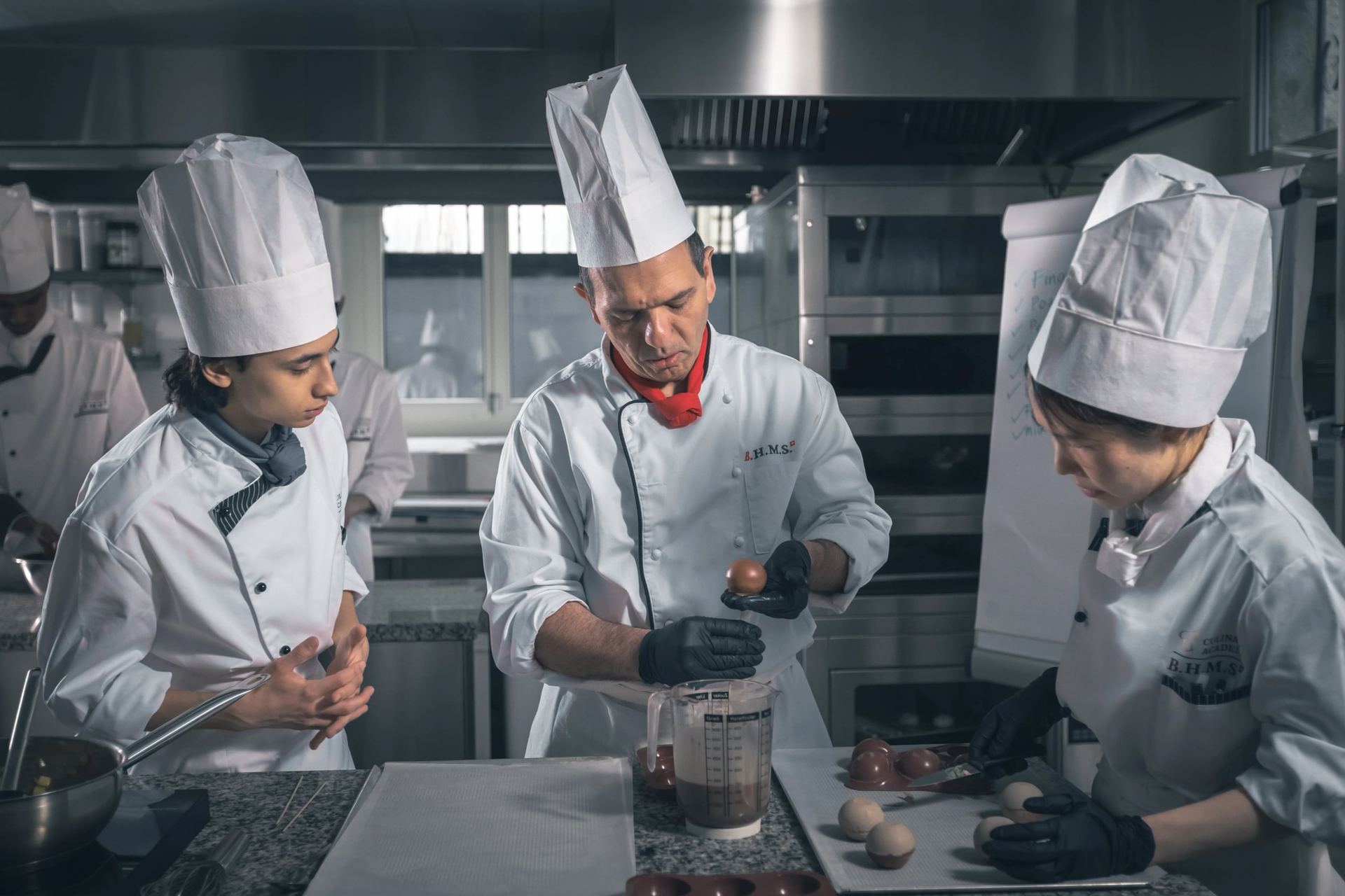 Chef in a B.H.M.S. jacket holds a chocolate sphere over melted chocolate as two students work beside him