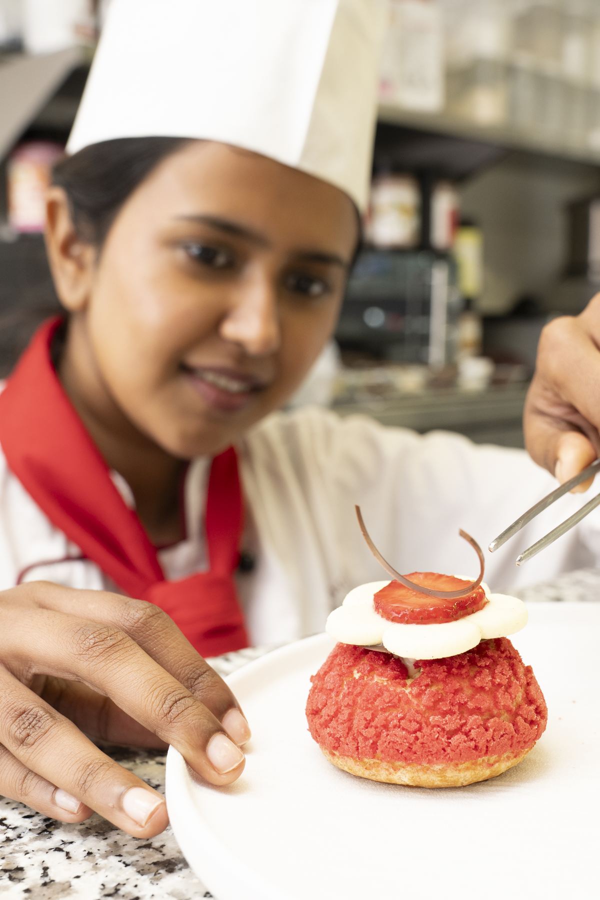 Pastry student with tweezers finishing a red craquelin choux bun topped with cream, strawberry and a chocolate arc