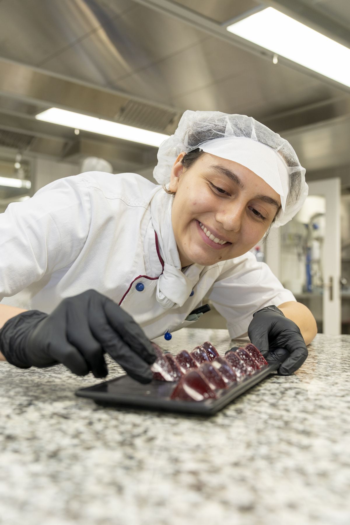 Pastry student in chef jacket and hairnet arranging glossy dark red confections on a black tray