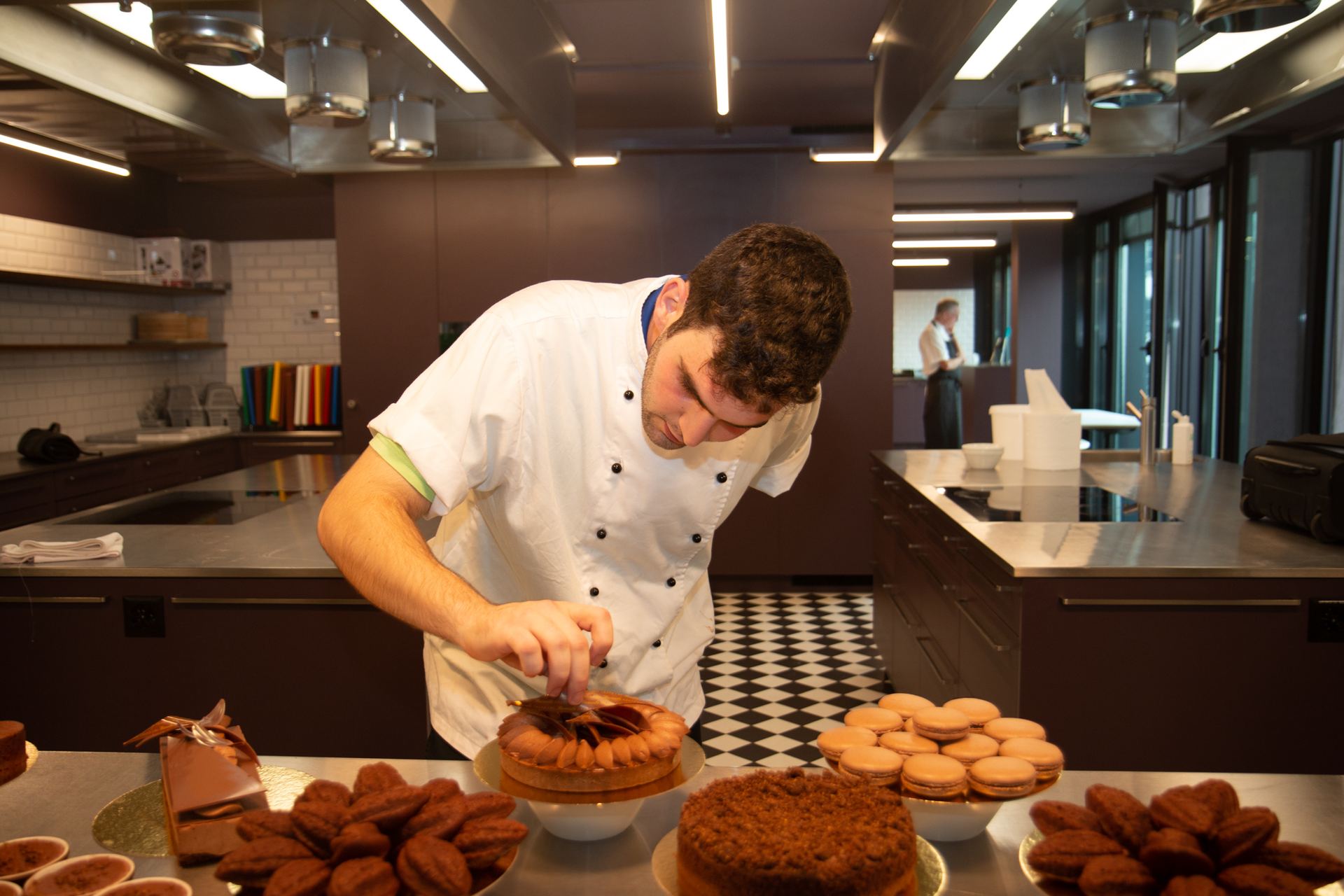 Pastry chef places a chocolate decoration on a tart beside macarons, madeleines and cakes