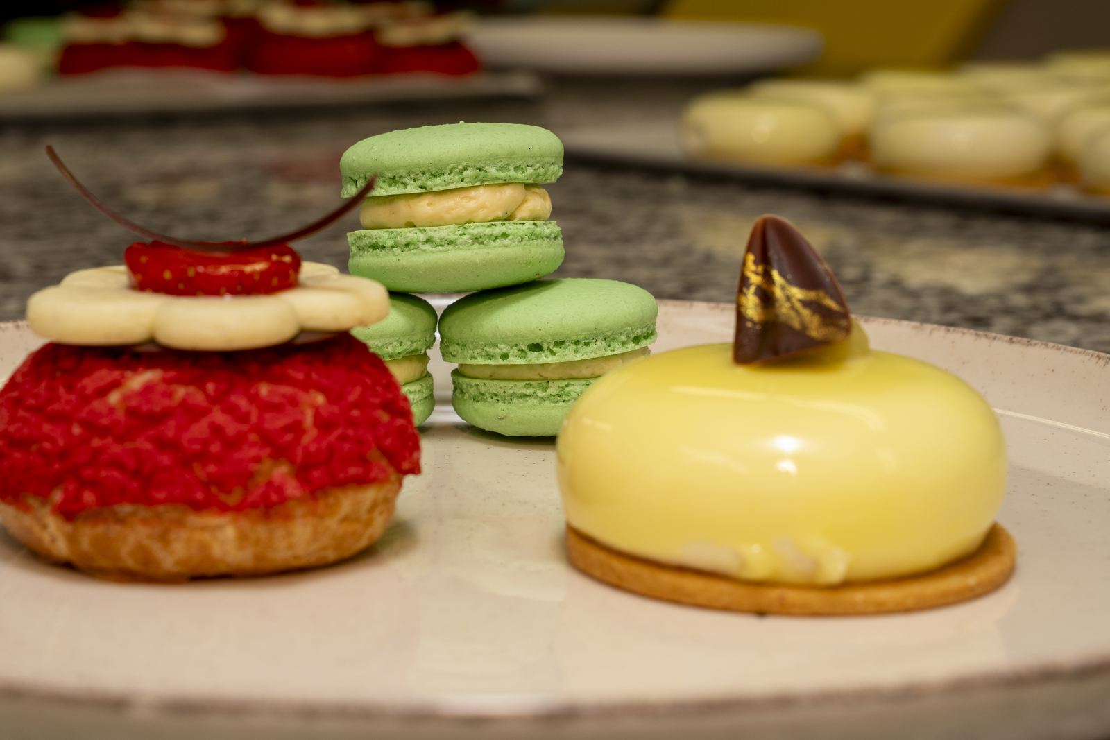 Red craquelin choux with strawberry, green macarons and a yellow mirror-glazed entremets on a pale plate