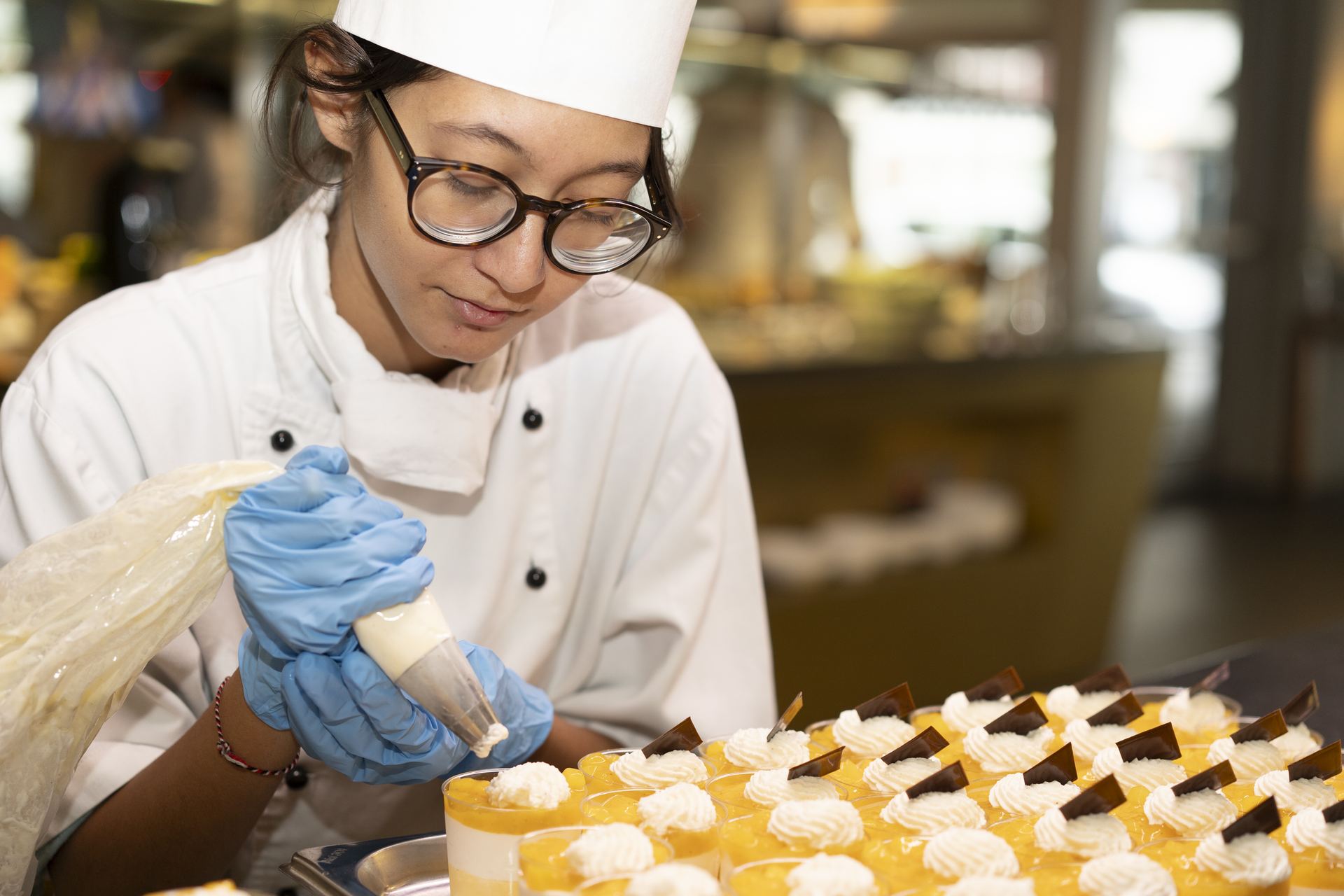 Pastry student in chef whites pipes cream onto a tray of layered fruit verrines with chocolate triangles