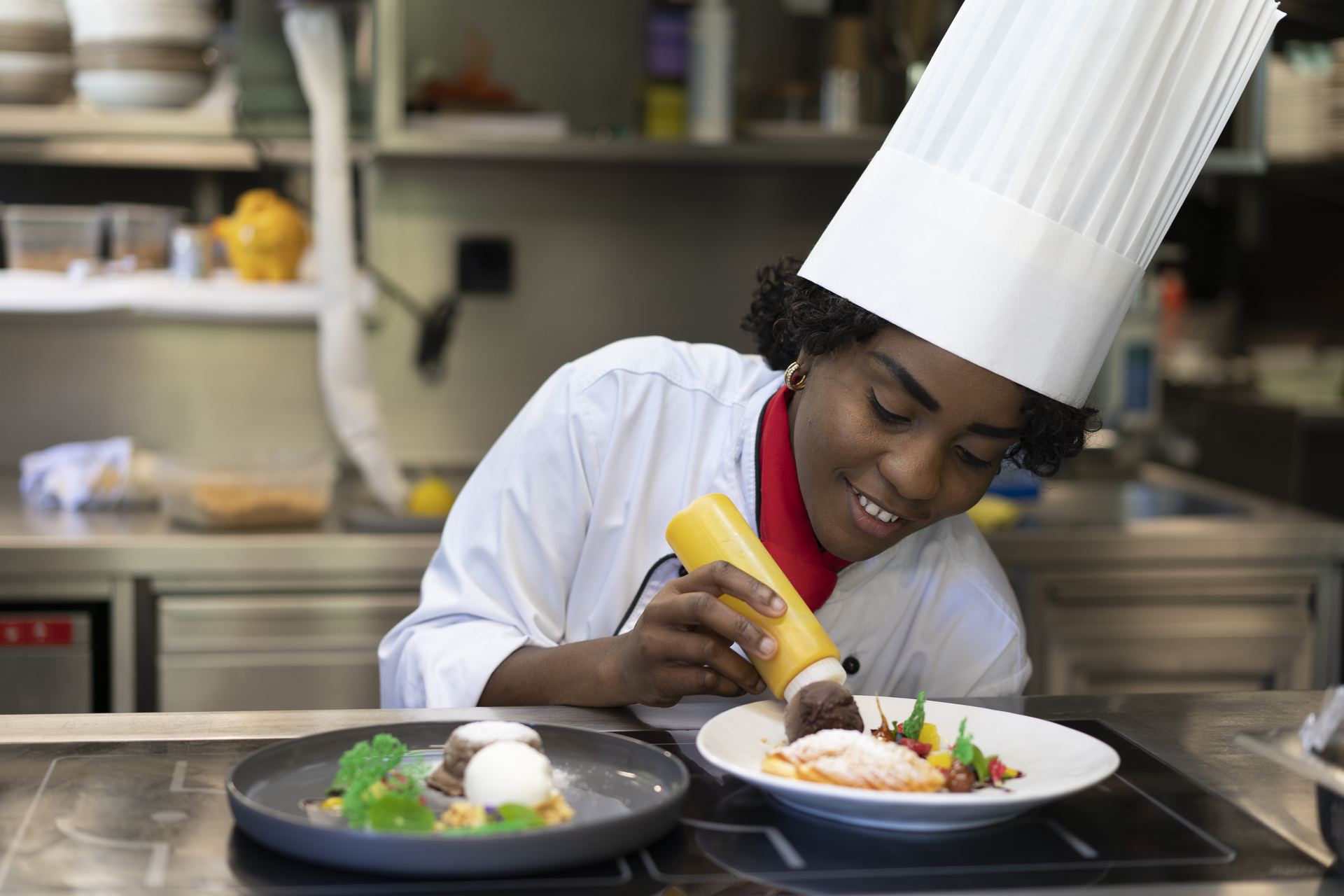 BHMS student in a tall chef's toque finishing a plated dessert with a squeeze bottle