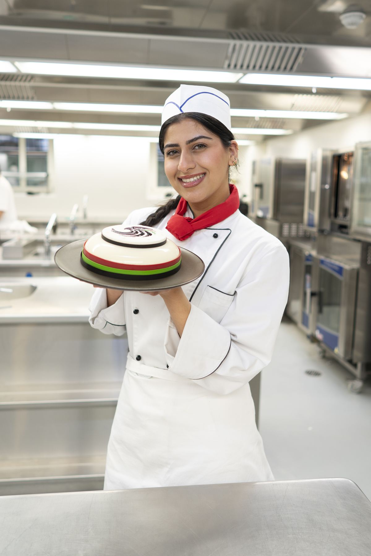 Smiling student in chef whites holds a domed cake with red, green and white bands in a teaching kitchen