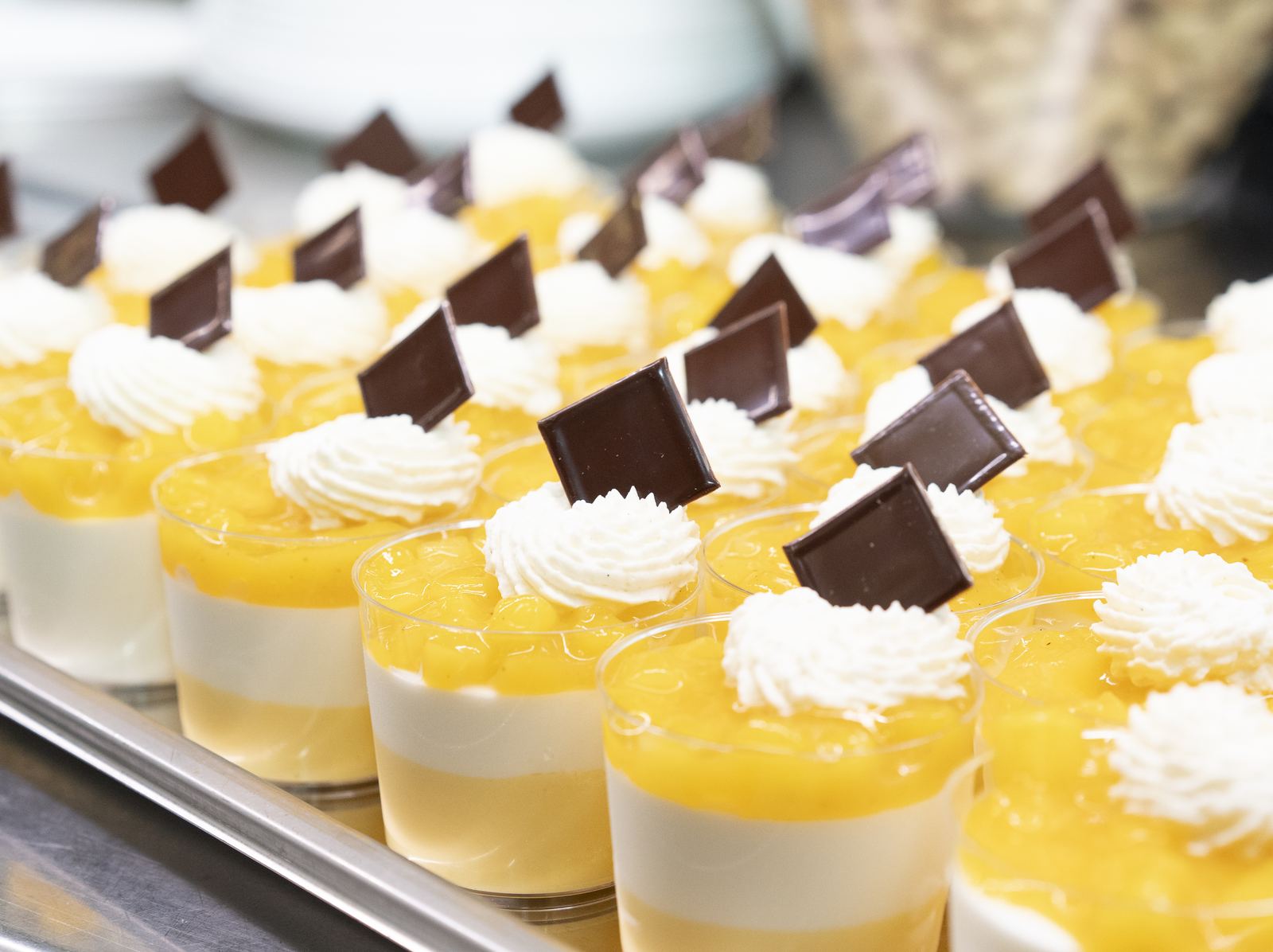 Rows of layered fruit and cream desserts in clear cups, each topped with piped cream and dark chocolate